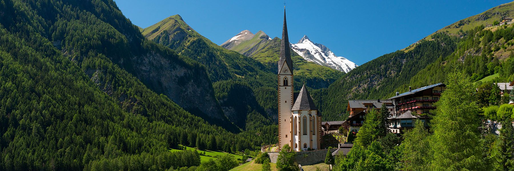 Ansicht auf ein kleines Bergdorf im Fokus die imposante Kirche-rechts davon ein Paar Häuser und ein Uriges Hotel- links und rechts erstrecken sich die mit grünen Bäumen verzierten Berge